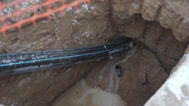 Mavi çizgili siyah kauçuk boru toprak çukura giriyor. Sert toprak duvarları, devam eden yeraltı altyapı çalışmalarını gösteriyor.