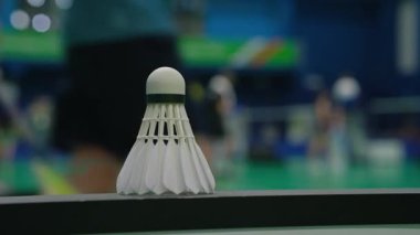 Shuttlecock düz zemin üzerinde dururken arka planda oyuncular belirdi. Kapalı alanda badminton sahası oyun anını öneriyor