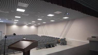 Koyu podyumlu sahne, düzgün sıra sıra gri sandalyelere bakıyor. Izgara tavan ve pastel duvarlar düzen ve sakin konferans ortamını vurguluyor