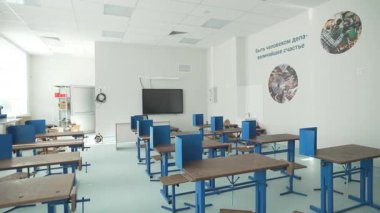 Sınıf, tahta masalar ve metal sandalyelerle boş kalıyor. Beyaz duvarlar motivasyon sloganı ve öğrenciler için yuvarlak eğitim posterleri gösteriyor