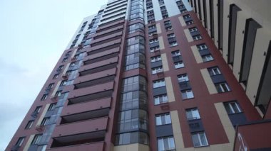 Cam balkonlu yüksek binalar alçak açılı çekimde temiz gökyüzüne karşı yükselir. Konut kompleksi içinde koyu kırmızı cepheli modern bina