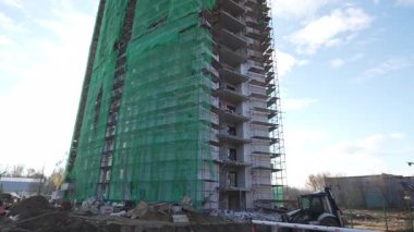 Bitmemiş gökdelen, gökyüzünün altında çırpınan yeşil muşambalara sarılı duruyor. Çelik kirişler ve beton levhalar üst katlarda açıkta duruyor.