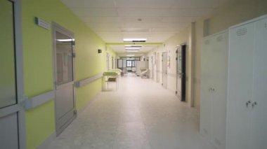 Geniş hastane koridoru temiz ve düzenli görünüyor. Işık cilalanmış fayanslardan yansıyor. Sakin tasarım tıbbi atmosferi destekler