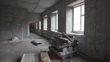 Parlak plastik pencereli, tamamlanmamış geniş bir oda. Dağınık yapı araçları ve tozlu köşelerde yığılmış malzemeler çıplak duvarlar arasında