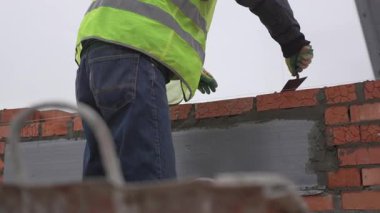 İşçi, çimento katmanını hızlı bir elle uygular. Tuğla yukarıda bekliyor ve her katman yükseklik ekledikçe temel yükseliyor.