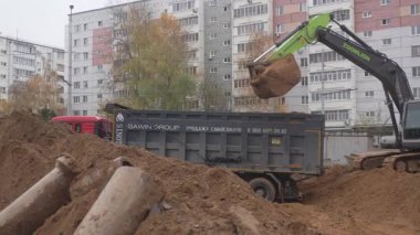 ALMATY, KAZAKHSTAN - 17 Ekim 2024: Modern kazıcı çok katlı binalara karşı kamyon römorkuna kuru toprak yükler. Toprak nakliyecisi ve kamyoncu kasabadaki çukura pislikle çalışıyor.
