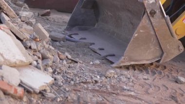 Ağır kazıcı kovası tozlu zeminde eski duvarların yanında enkaz yığınına doğru sürünüyor. Yavaş kova moloz yığınına doğru sürekli hareket ediyor.