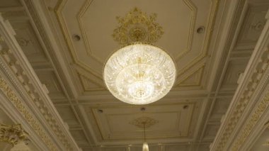 Geometrik desenlerle dekore edilmiş tavan büyük avizeyi çerçeveler. Tavan süslemelerinin simetrisi ışıkla parlayan avizeyi sarar