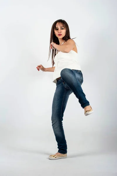 Full length portrait of a young woman dancing hip-hop against white background in a kicking position with one foot up in the air