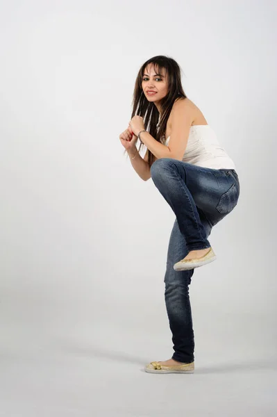 Young beautiful brunette woman in jeans and sneakers on a white background in a kicking position with one foot up in the air