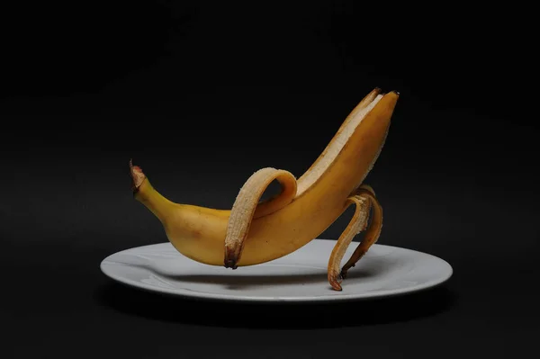 半剥皮香蕉站在空气中的瓷盘上 底座是黑色的 — 图库照片