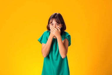 Korkak kız, elleri sarı arka planda, ağzı kapalı.