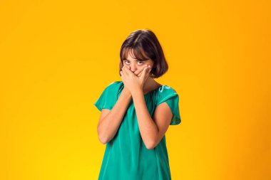 Korkak kız, elleri sarı arka planda, ağzı kapalı.