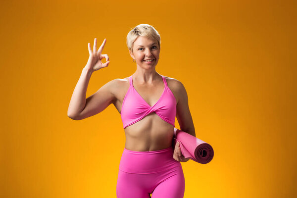 Fitness woman with short blond hair holding yoga mat and showing "ok" gesture isolated on yellow background.