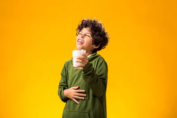 Lactose intolerance. Dairy intolerant unhappy child boy holding glass of milk and feeling abdominal pain over yellow background.