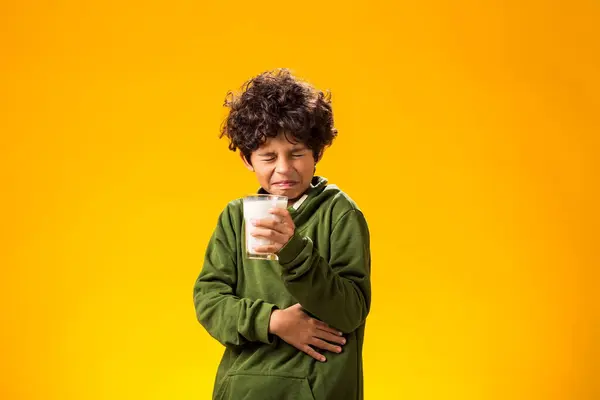 Lactose intolerance. Dairy intolerant unhappy child boy holding glass of milk and feeling abdominal pain over yellow background.