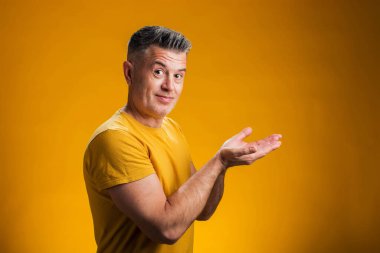 Neşeli bir şekilde gülümseyen bir adamın portresi. Avuç içiyle göstererek sarı arka plandaki kameraya bakıyor. Reklam kavramı