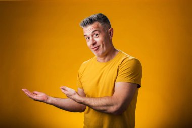 Neşeli bir şekilde gülümseyen bir adamın portresi. Avuç içiyle göstererek sarı arka plandaki kameraya bakıyor. Reklam kavramı