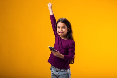 Sosyal medyada cep telefonuyla mesaj atan, mobil ağ hizmetlerini kullanan, internette sohbet eden küçük bir kızın portresi. Sarı arka planda kapalı stüdyo çekimleri.