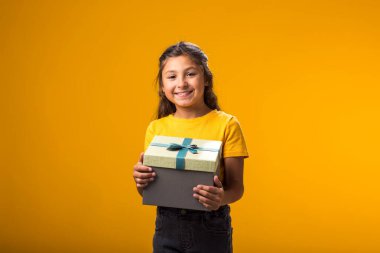 Sarı arka planda hediye kutusunu tutan gülümseyen kız portresi. Doğum günü ve kutlama konsepti