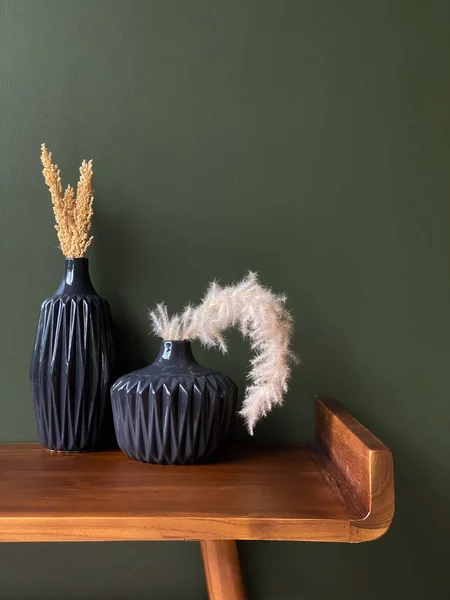 Two black vases with dried grass and plants on a wooden table against a green wall