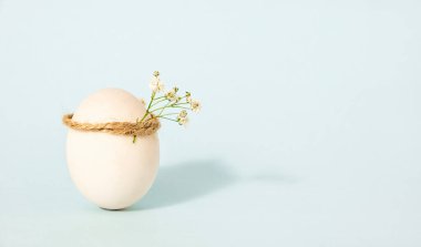 Single white chicken egg with improvised wreath on pastel blue background as a Easter holiday concept, copy space 