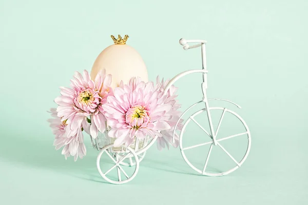 ピンクの菊の花と小さな王冠と単一の鶏の卵と白い三輪車の小さな置物 コピースペースとイースターの概念 — ストック写真