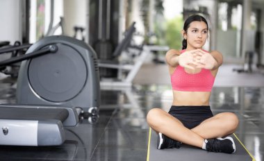 Genç bir kadın geniş bir spor salonunda esneme hareketleri yapıyor.