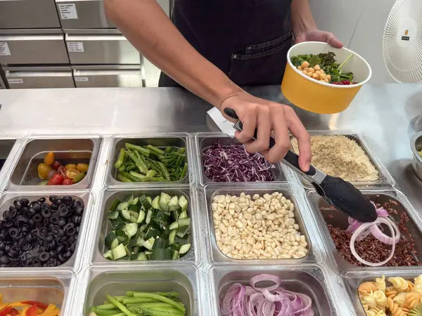 Penang, Malezya - 26 Nisan 2025: Bir el salata barından salatalık, soğan, zeytin ve çeşitli sebzeler içeren taze malzemeler seçerken görülüyor..