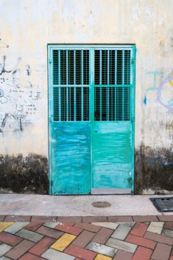 Penang, Malezya 23 Temmuz 2025 yıpranmış bir turkuaz çift kapılı ve desenli bir ızgara solmuş bir duvara karşı duruyor ve tarihi bölgenin hareketli sokak manzarasının cazibesini arttırıyor..