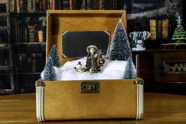 Christmas scene in vintage suitcase with snowman and young girl figurine with old library background