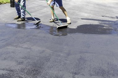 İki adam güneşli bir günde asfalt yola çalı çırpı ve cam sileceğiyle mühür basıyor.