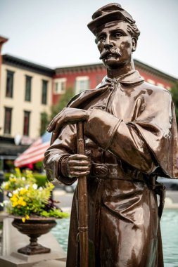 Güneşli bir günde arka planda Amerikan bayrağıyla kasaba merkezinde nöbet tutan dikey bronz savaş askeri heykeli.