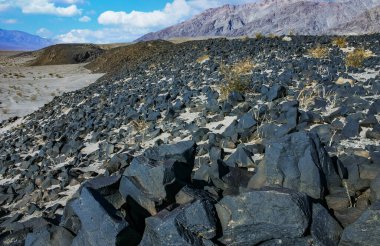 Ölüm Vadisi Ulusal Parkı 'ndaki siyah lav kayalarının geniş bir görüntüsü. Kayalar yere saçılmış, bazı küçük kum parçalarıyla görülebilir. Mavi gökyüzü beyaz kabarık