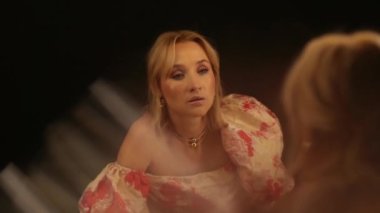 Açık renk elbiseli güzel bir kadın karanlık bir soyunma odasında aynanın yanında duruyor. Üzüntü, yorgunluk ve kendine şefkat gösteriyor..