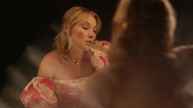 Açık renk elbiseli güzel bir kadın karanlık bir soyunma odasında aynanın yanında duruyor. Üzüntü, yorgunluk ve kendine şefkat gösteriyor..