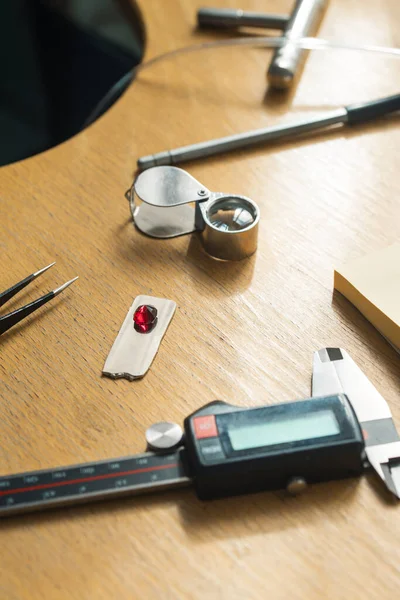 stock image Jewelry craftsmanship. Jeweler tweezers, Magnifier, Ruby gemstone. The jeweller engraver tools on wooden vintage desk.