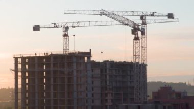 Bir Konut Binası İnşaatı. Zaman atlaması. İnşaat halindeki bir tesisin üzerinde üç vinç kulesi var. Akşam gökyüzünün arka planına karşı yoğun çalışma devam ediyor