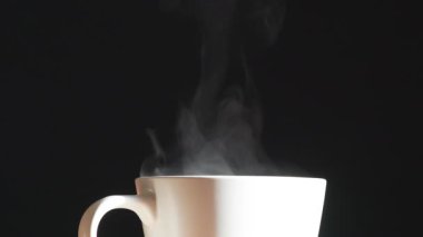 Sıcak Sütlü Beyaz Kupa. Bir fincan sıcak kahveden ince bir buhar damlası yükselir ve zarafetle döner. Arkaplan ışığı siyah arkaplanda fincanın bej rengini aydınlatır