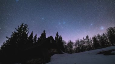 Astronomy time lapse of Starry night sky and stars motion fast above wooden hut in wilderness forest nature in winter snowy landscape. Timelapse, Zoom in, Tilt up. 
