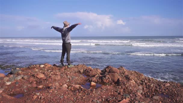 戴帽子的人想象着 在摩洛哥的非洲自然景观中 梦想着像鸟儿一样在岩石沙滩上飞越大西洋水面 暑期旅行自由探险 — 图库视频影像