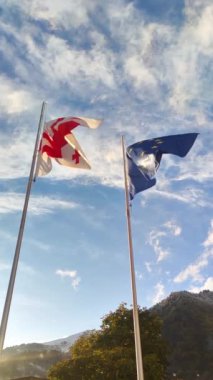 Gürcistan ve Avrupa Birliği 'nin bayrağının güneşli bir günde mavi gökyüzüne doğru esen rüzgarda dalgalandığı dikey video, düşük açı yavaş çekim.