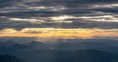 Dramatik bulut katmanlarıyla nefes kesici altın gün batımı gökyüzü ve yuvarlanan tepelerin ve dağların üzerinde parlayan güneş ışınları. Sinematik projelerde kullanılmak üzere çarpıcı bir zaman atlaması ile yakalanan doğa