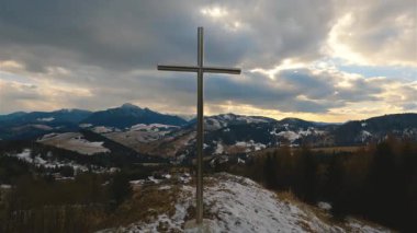 Paslanmaz çelik haç, karlı dağ manzarası ve ılık günbatımı ışığıyla bir tepenin üzerinde duruyor. İlkbaharın başında ruhani, huzurlu doğa manzarası. 