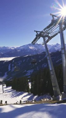Zell am See kayak merkezindeki teleferik inşaatının dikey manzarası. Açık mavi gökyüzü, karlı yamaçlar ve dağlık tepelerde güneş patlaması. Kış yolculuğu, turizm ve eğlence amaçlı görüntüler için ideal.