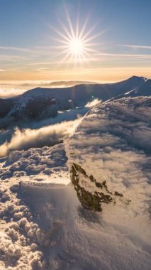 Gün batımında karlı dağlarda güzel bir dikey zaman atlaması. Altın ışık sisli tepelerde parlıyor, bulutlar vadilerde sürükleniyor, ve bu büyülü donmuş doğa sahnesinde kar parıldıyor.