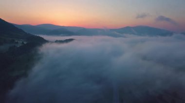 Güneş doğarken dağın tepelerinde yuvarlanan sisin dingin güzelliğini gösteren hava manzarası. Mavi saatin yumuşak ışığı, sakin sabah atmosferini güçlendirir..