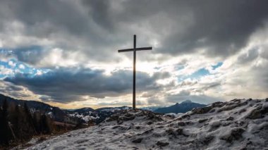 Akşam bulutları dramatik bir gökyüzünde ilerlerken karlı dağ doğasında çelik bir haç. Kış mevsiminin bu zaman aşımı inancı, umudu ve maneviyatı çağrıştırıyor. Mükemmel dini görüntüler