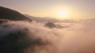 Puslu orman doğası ve altın gündoğumu ışığında dağların insansız hava görüntüsü. Sis yavaşça dağların üzerinde sürükleniyor, nefes kesici kırsal alan ve büyülü sabah atmosferi yaratıyor..