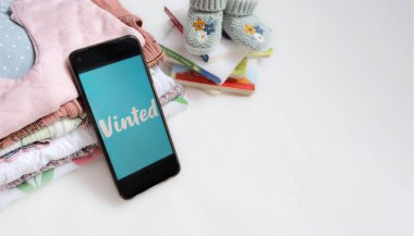 Braunschweig, Almanya - Juli 03, 2023: Mobil uygulamalı akıllı telefon Vinted ve çocuk kıyafetleri, kopyalanmış beyaz arkaplan üzerine kitaplar. İnternet üzerinden çocukların ikinci el giysilerinin satışı